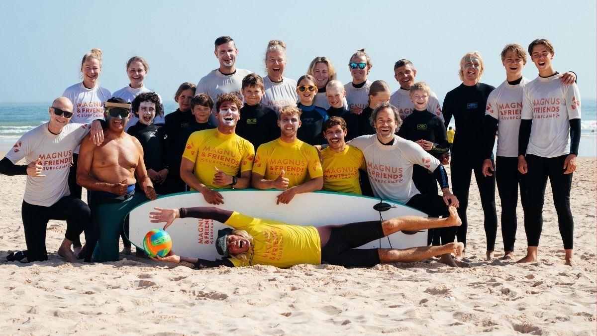 En stor surfgrupp med deltagare i olika åldrar som står och poserar glatt på stranden ihop med surfinstruktörerna