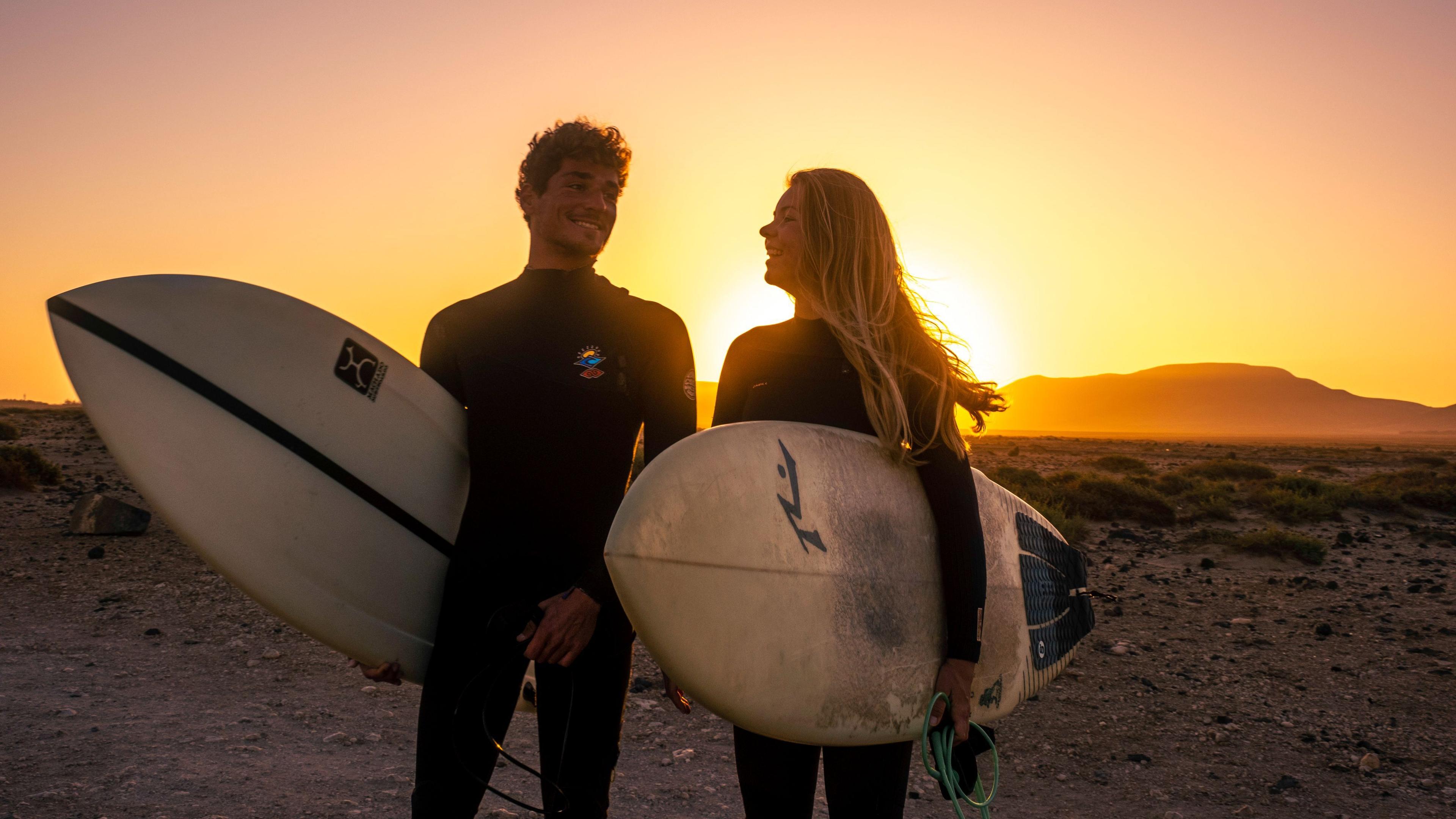 Två surfare som står med sina surfbrädor framför en magisk solnedgång