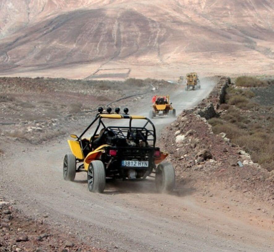 Dune buggies kör längs en dammig grusväg med ökenlandskap och berg i bakgrunden