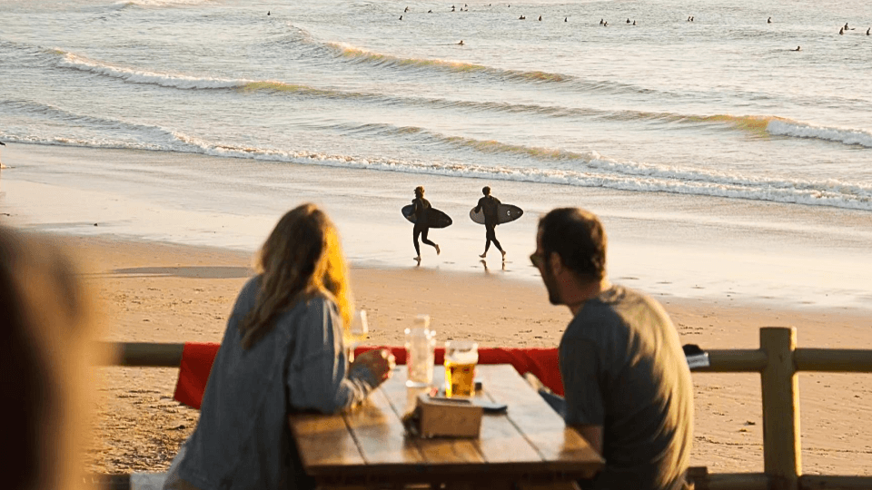 Två personer njuter av en drink vid ett bord med utsikt över stranden, där surfare med brädor går längs vattnet i solnedgången