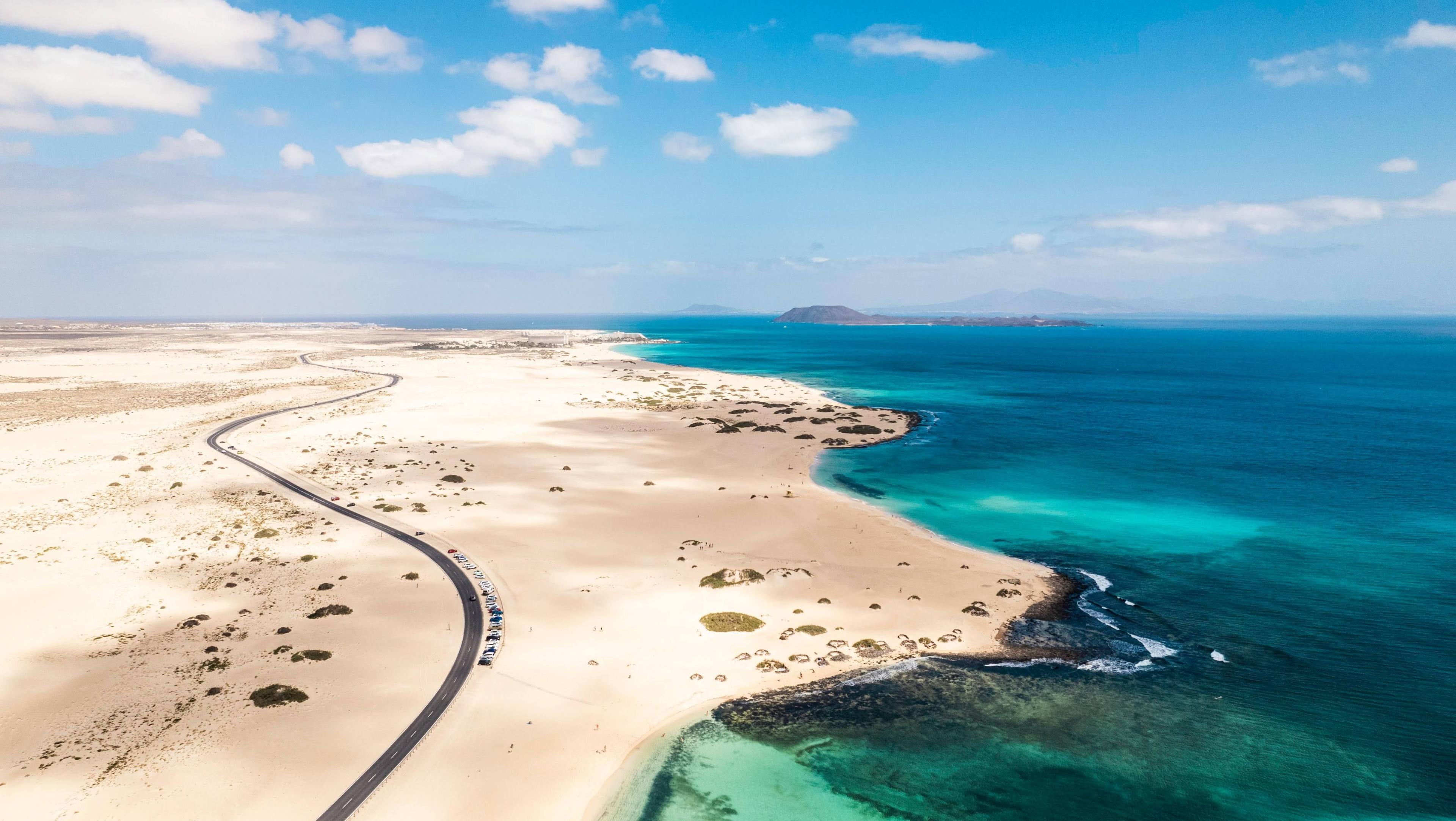 Flygbild över Fuerteventura med slingrande väg, sanddyner och turkost hav.