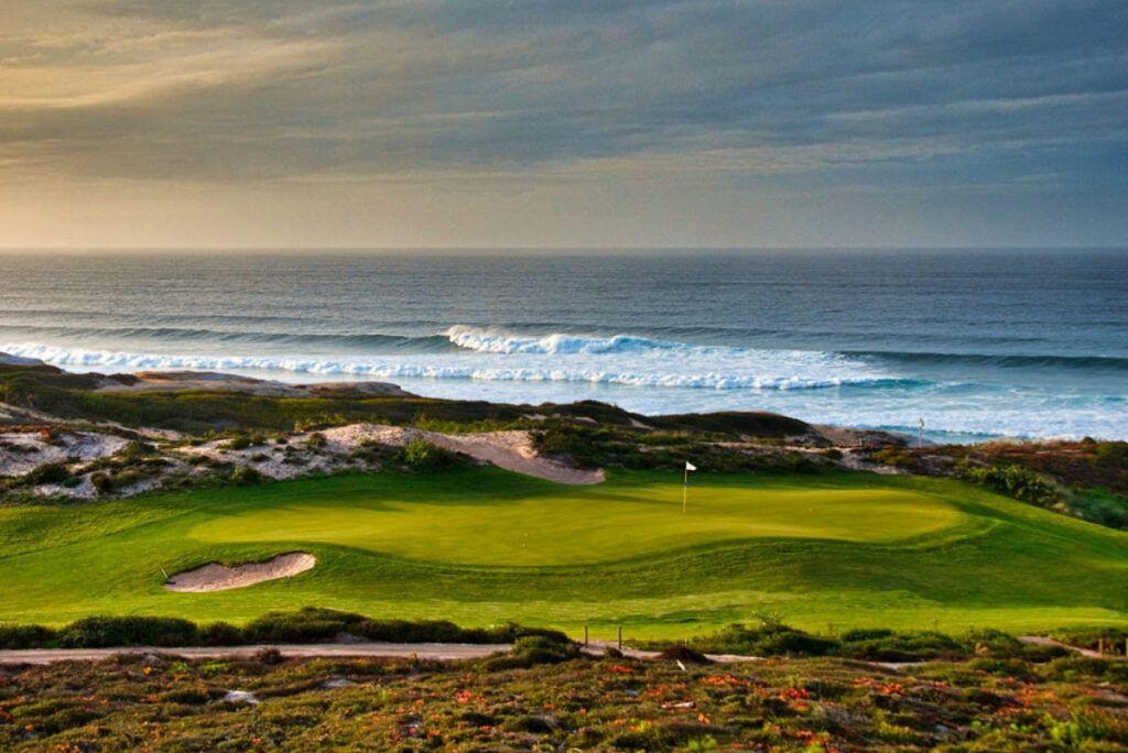 En grön golfbana med utsikt över havet och stora vågor, omgiven av sanddyner och vacker natur