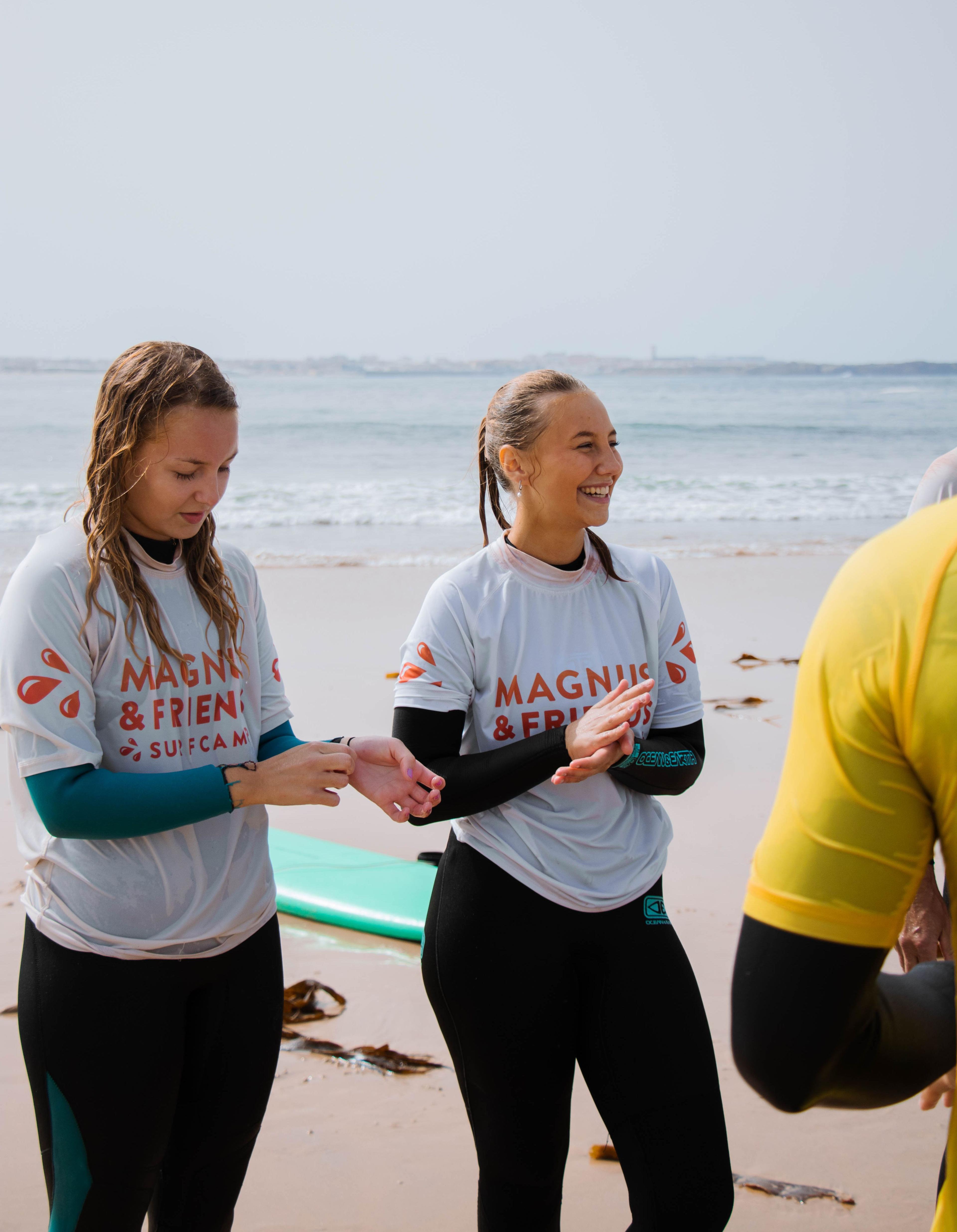 Två kvinnor i Magnus & Friends-tröjor får personlig surfcoachingsession på stranden, med havet i bakgrunden