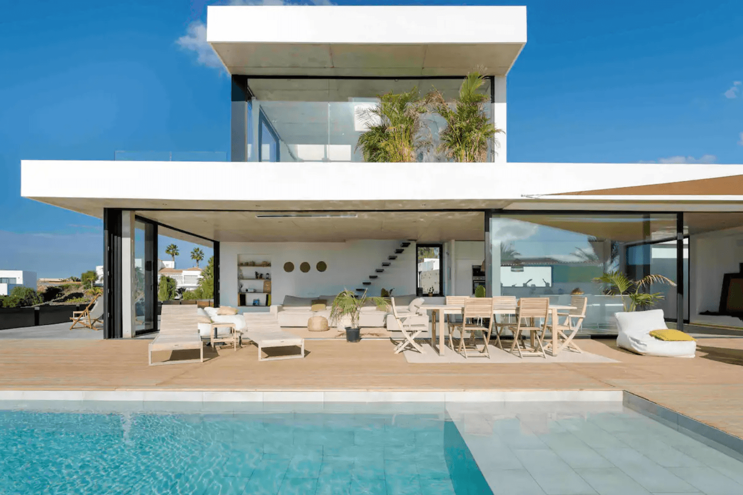 Modern villa med öppna ytor, solstolar och matplats på terrassen, samt en pool under klarblå himmel.