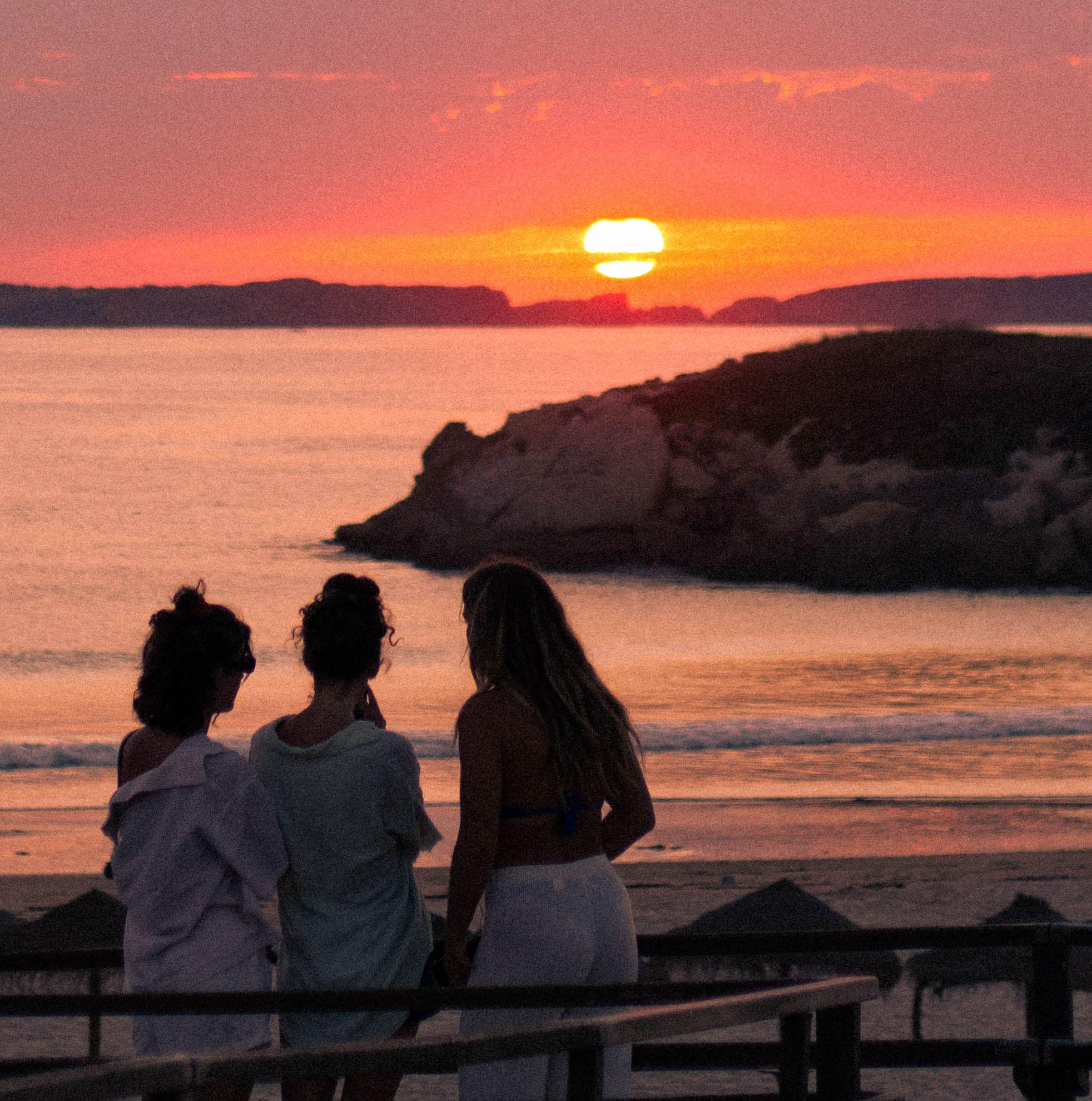 Tre vänner står på en träbrygga och blickar ut över havet under en magisk solnedgång