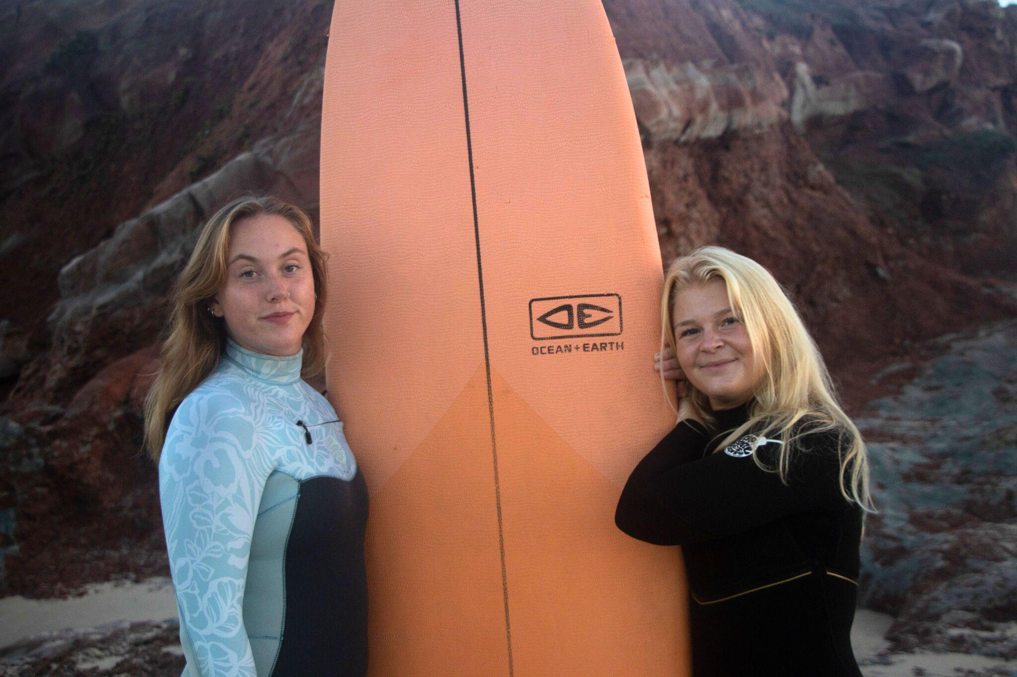 2 tjejer som står lutandes mot en orange surfbräda framför röda klippor