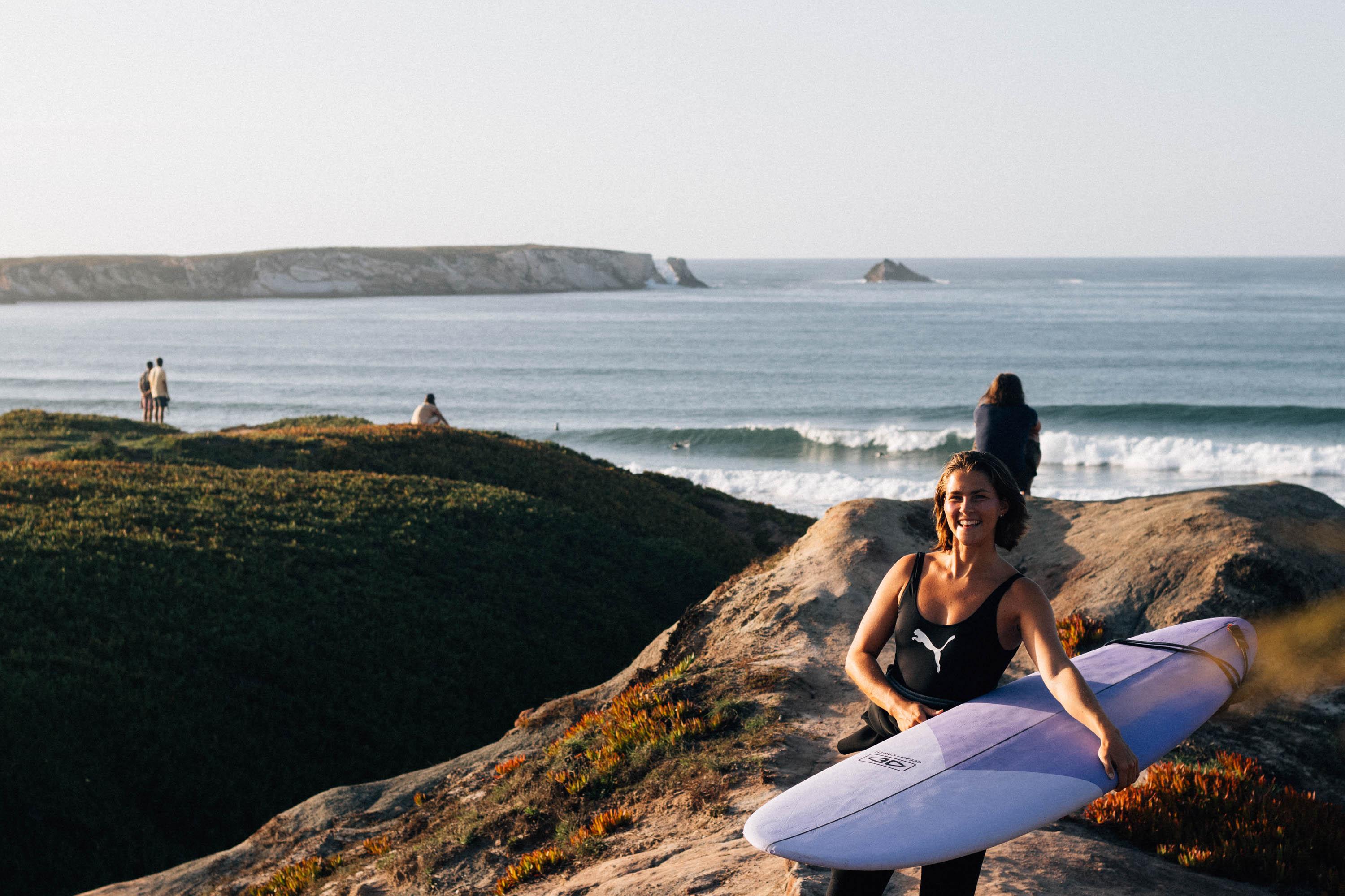 Surfare med bräda på en klippa vid havet i Baleal, med vågor och solig kust i bakgrunden