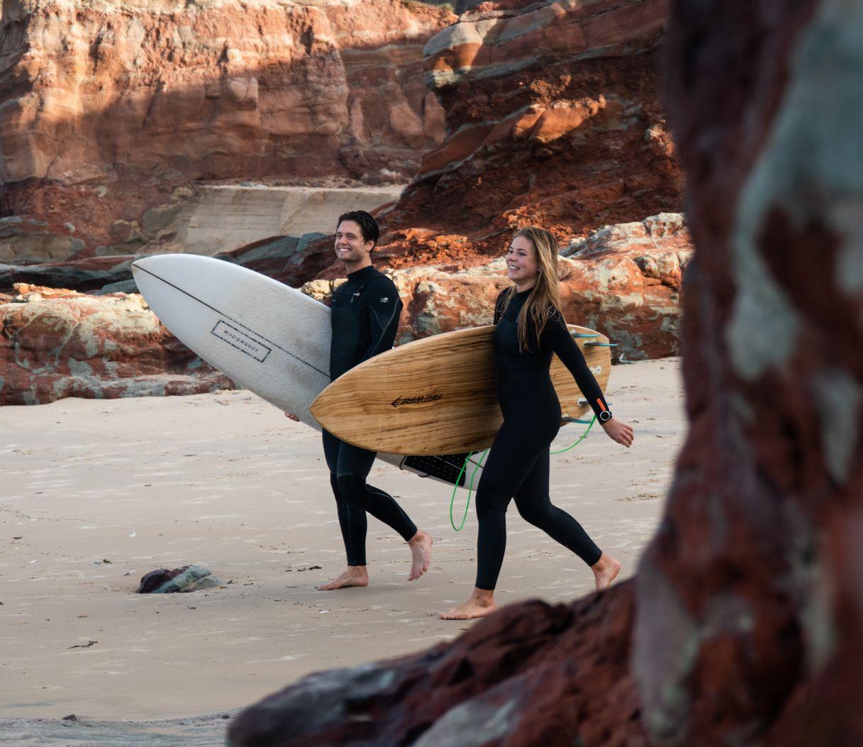 Två surfare på väg till stranden med sina brädor, omgivna av röda klippor och sand