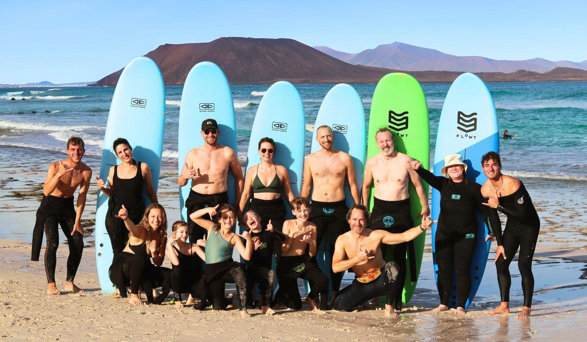 En surfgrupp poserar tillsammans på stranden framför surfbrädor, med havet och Fuerteventuras vulkaniska landskap i bakgrunden