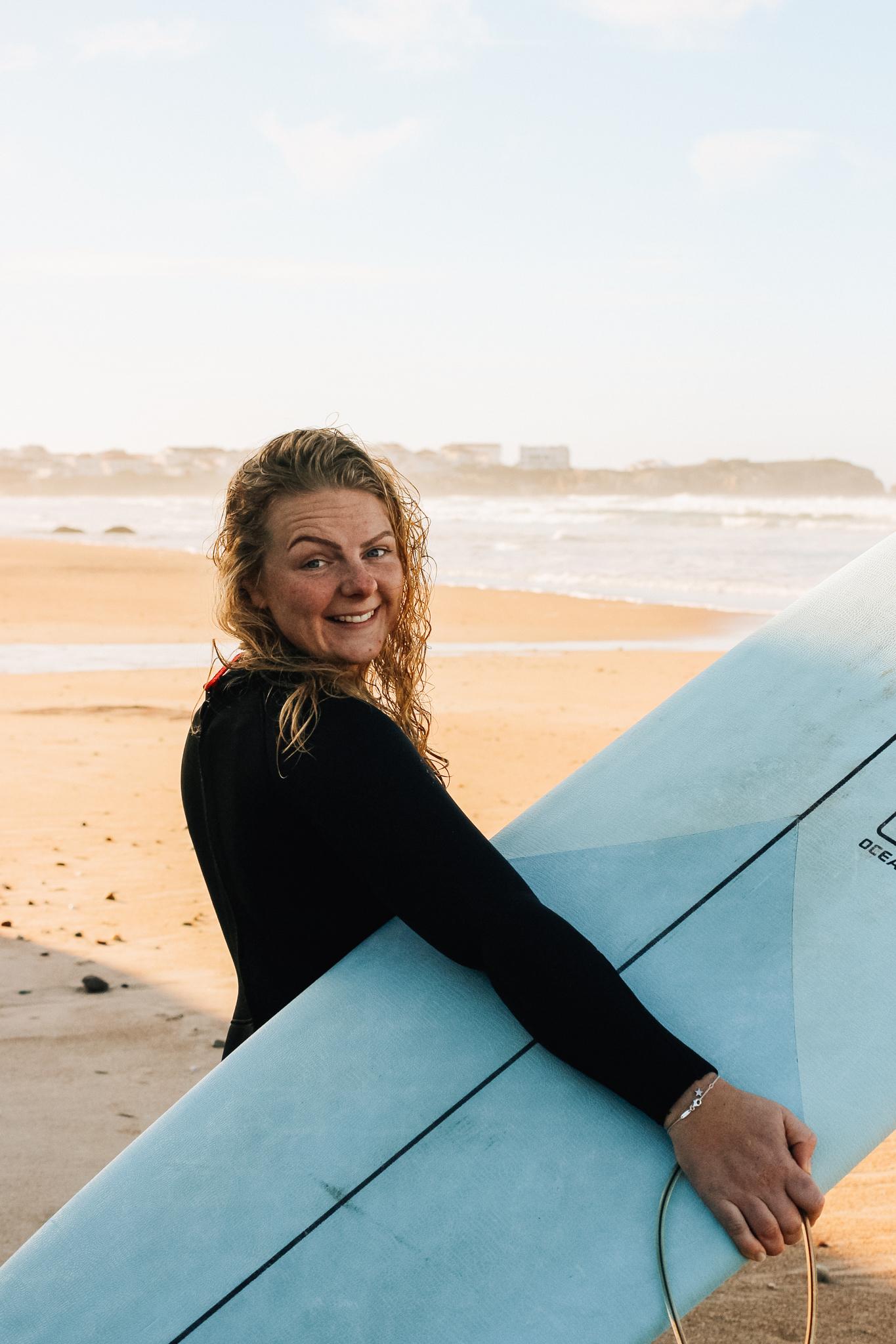 Alma står med en surfbräda på stranden och ser glad ut