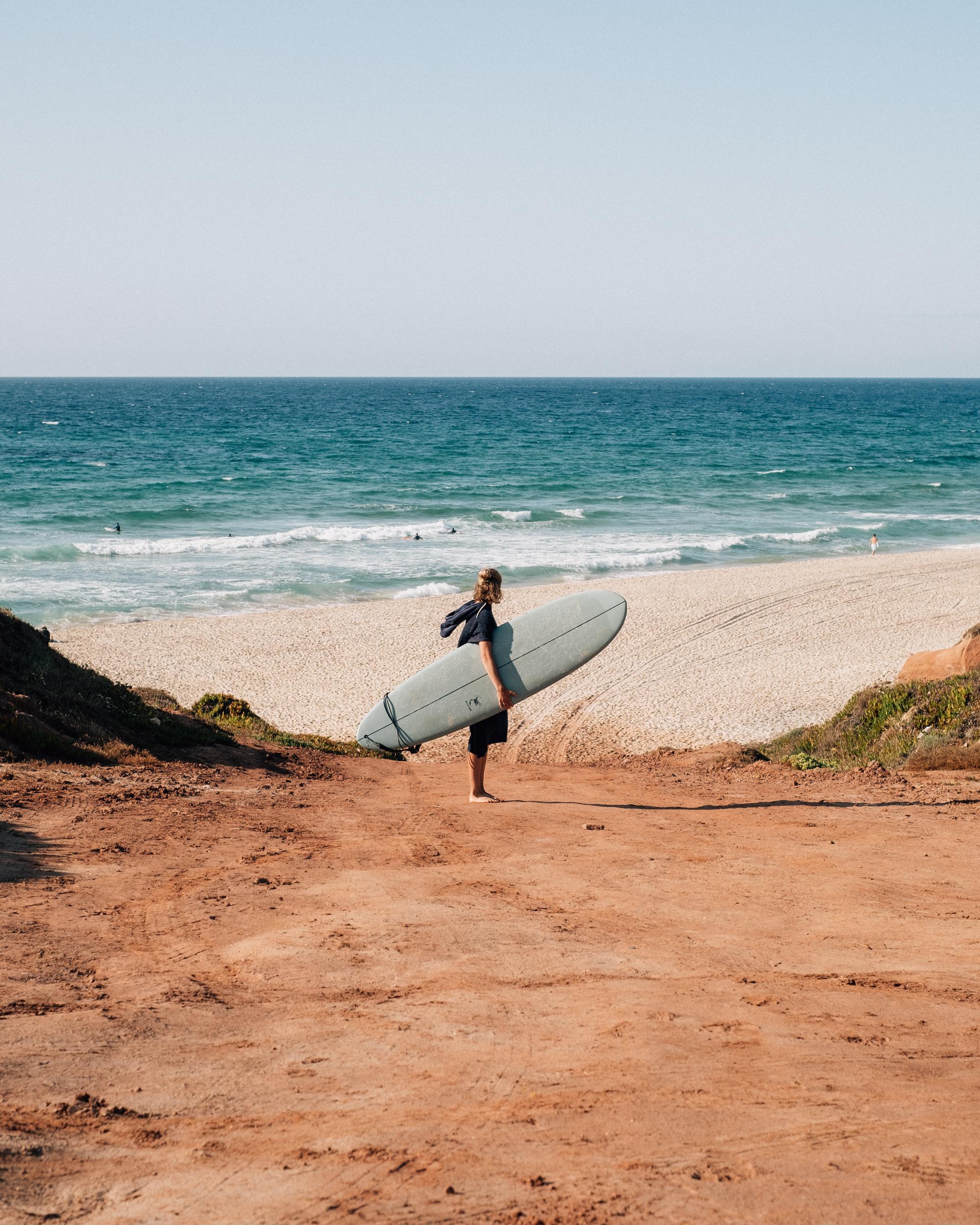 Surfare som står med en surfbräda nere vid stranden med havet i bakgrunden