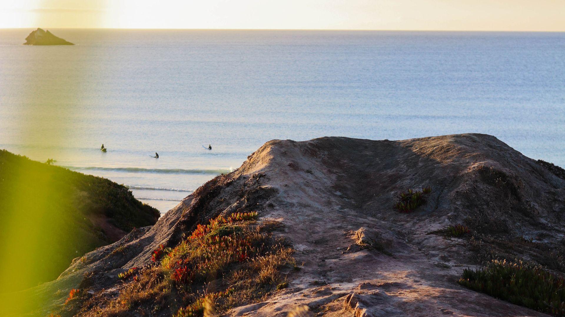 Surfare vid stranden i Portugal i solnedgången, fotograferat från klipporna ovanför stranden.
